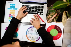 Person using a laptop with marketing charts, a red pen, and a small plant on the desk.